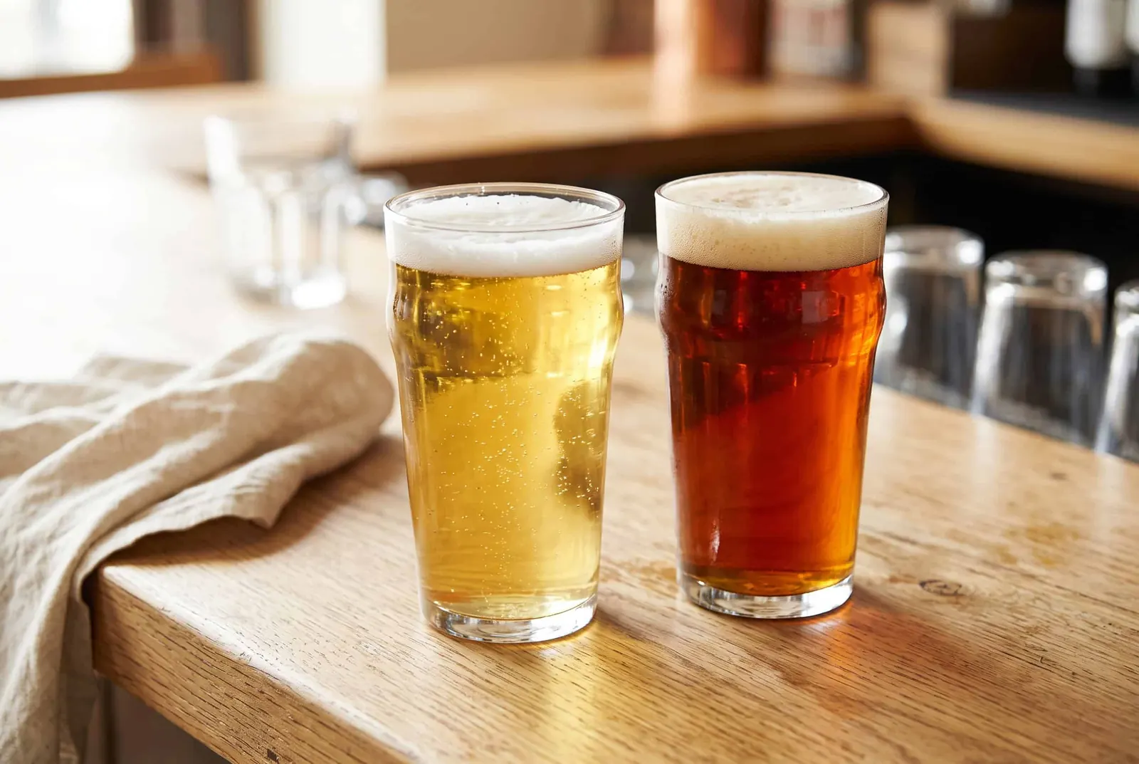 A pint glass of pale light lager beside an amber ale on a wooden bar